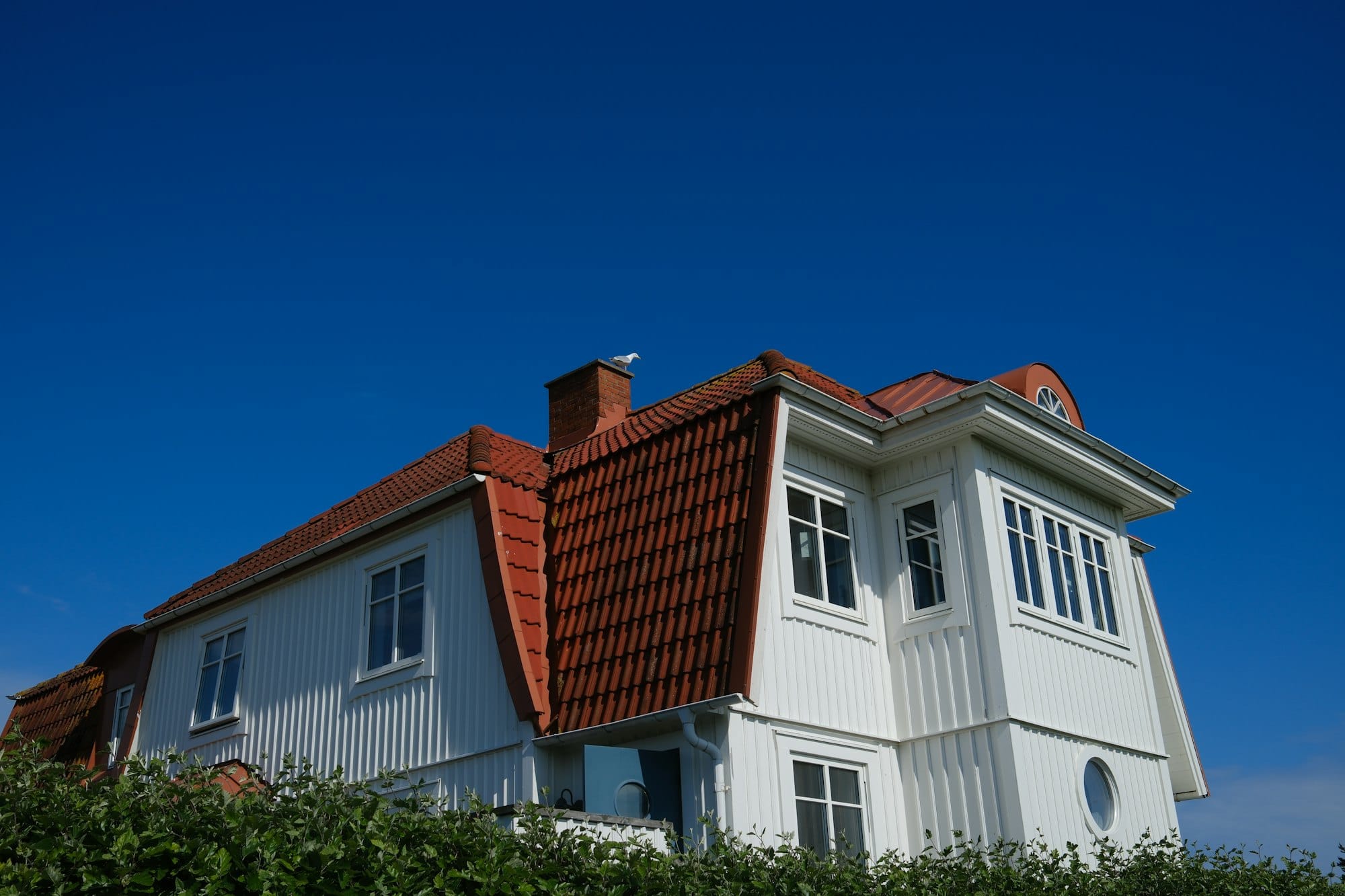 Hvid villa med rødt tegltag, tagrender og klippet hæk under en blå himmel